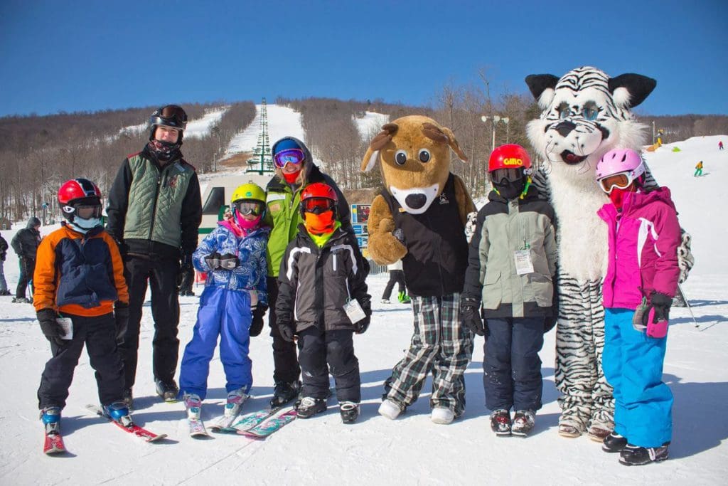 Several adults and kids in full ski gear stand with the two mascots at Whitetail Resort on a sunny winter day at the top of a slope.