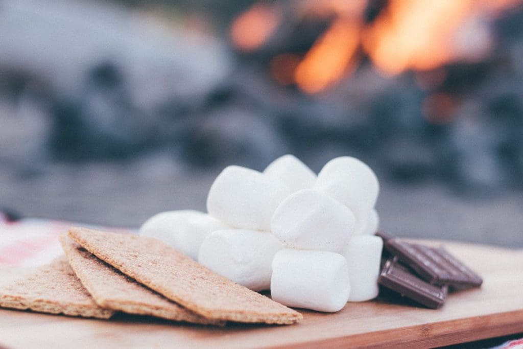 Graham crackers, marshmallows, and chocolate wait to become s'mores, with a fire blazing in the distance.