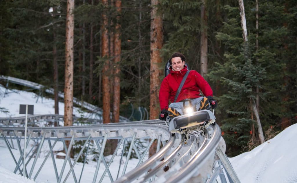 A man in a red coat smiles as he enjoys a ride down the alpine coaster, one of the best things to do when visiting Aspen during the winter with kids.