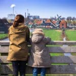 A mom and her child lean over a bridge looking at a beautiful Amsterdam scene, including greenery, a windmill, and buildings.