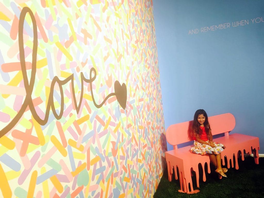A young girl sits on a bench at the Museum of Ice Cream that looks like a melted ice-cream cone next to a large sign that says "LOVE".
