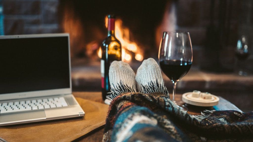 Feet stick out from a blanket with a view of a fireplace, wine, and computer, planning special surprises  is one of the best ways to add romance on a family road trip.