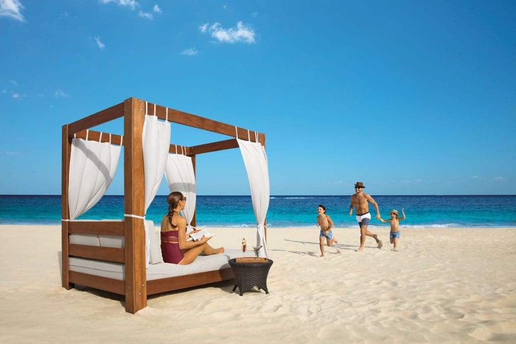A mom sits in a cabana, while dad plays with two kids on the beach at Dreams Riviera Cancun Resort & Spa.