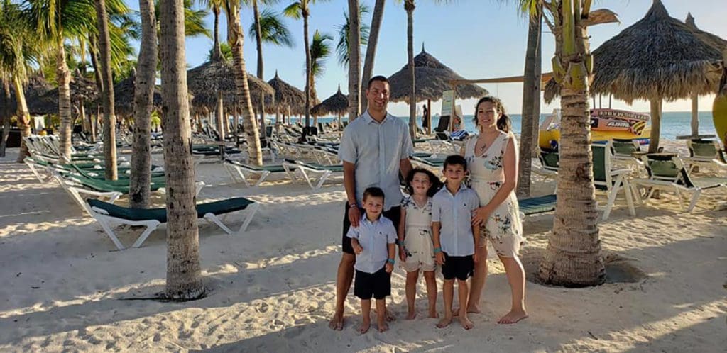 A family of fives stands together on Palm Beach in Aruba, with cabanas and the ocean in the background.