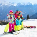 A mom and three kids, all in colorful snow gear, helmets, and skis, enjoy a sunny day on-mountain.