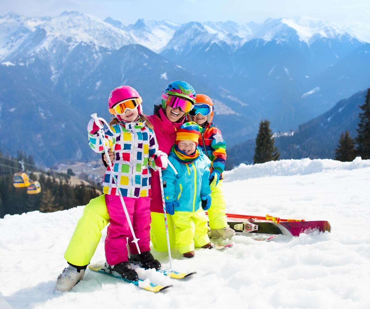 A mom and three kids, all in colorful snow gear, helmets, and skis, enjoy a sunny day on-mountain.