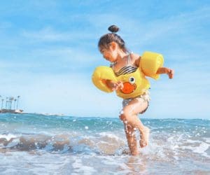 A young Asian child, wearing a duck-themed life jacket, spins in the ocean waves.