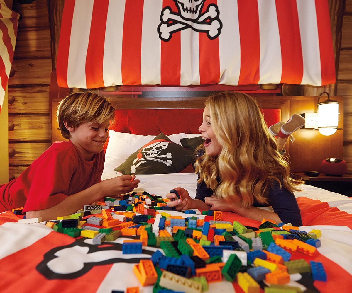 Two kids sit on a bed in a pirate-themed room looking at a large pile of legos.