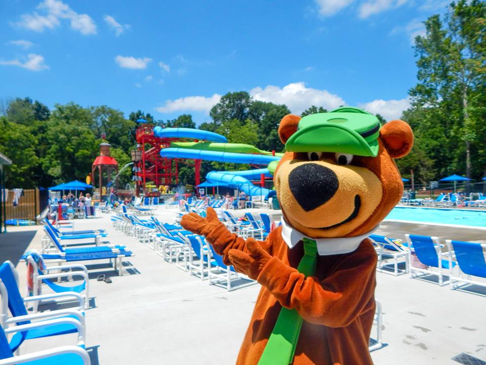 A Yogi Bear mascot points toward an outdoor waterslide area and pool deck at one of the Jellystone Parks in Maryland, one of the best themed hotels in the United States for families.
