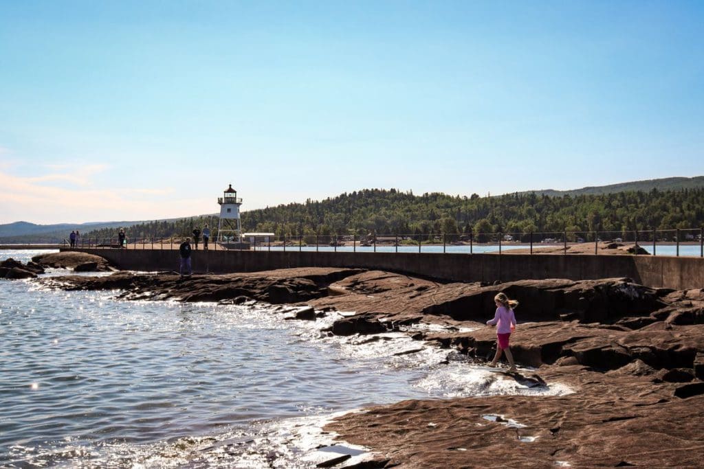 A young girl wanders about the large rocks and tide-pools near the Grand Marais Lighthouse, with the lighthouse and Sawtooth Mountains in the distance.