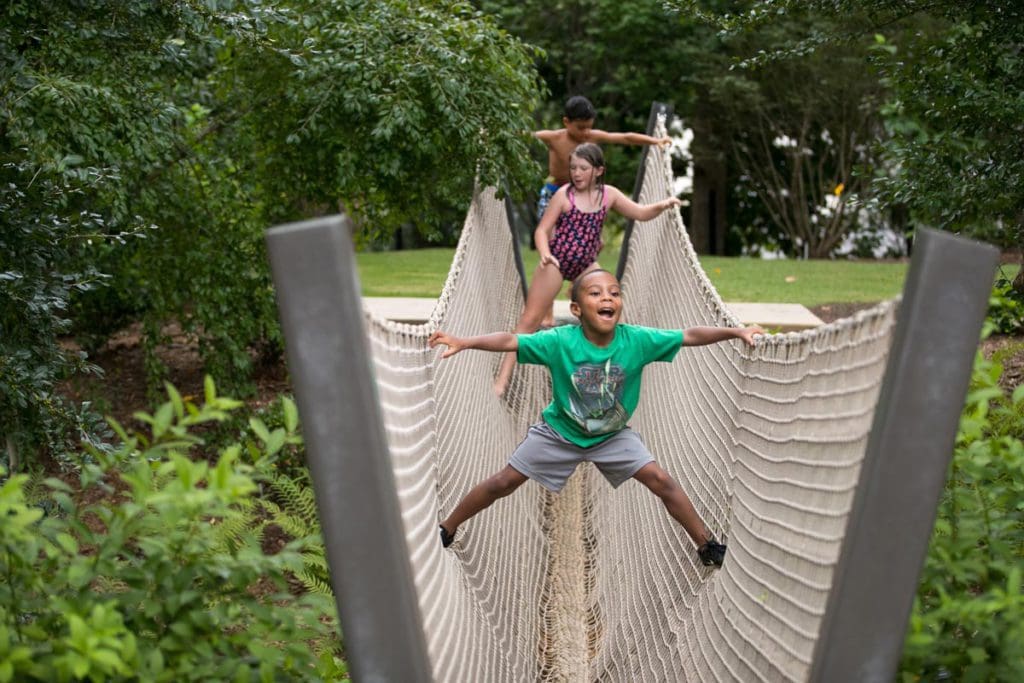 Three kids work their way across a large rope-style bridge at the Atlanta Botanical Garden.