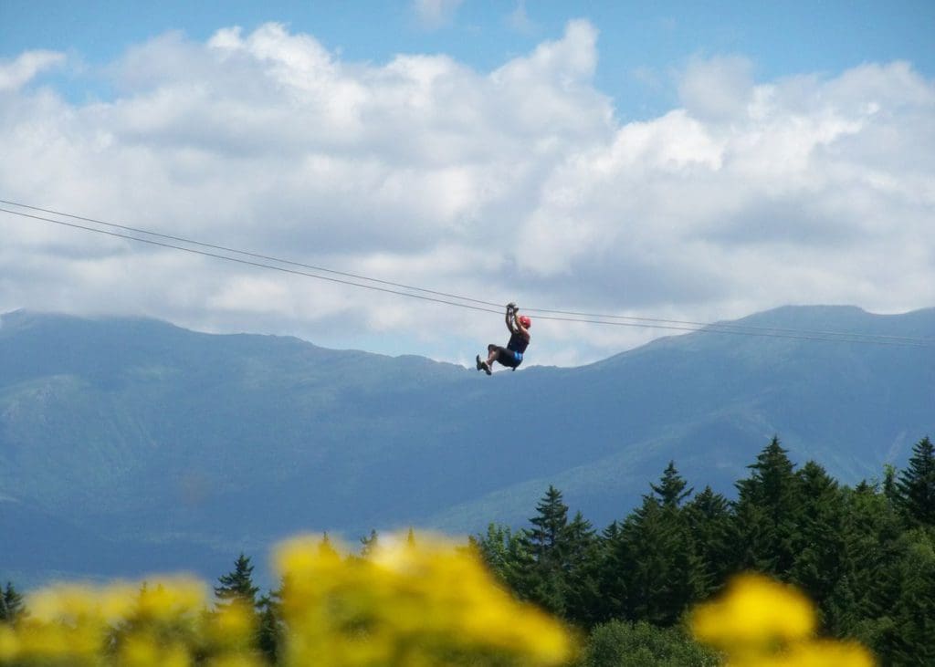 A woman zip-lines above the tree line, with the White Mountains of New Hampshire in the distance.