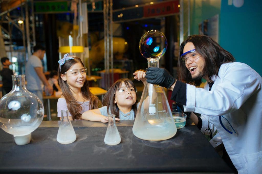 Two kids watch on as a staff member does a science experiment with beakers at DISCOVERY Children's Museum.