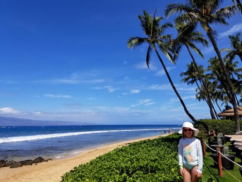 A young girl, wearing a swim suit and white beach hat, stands near a beach in Hawaii.
