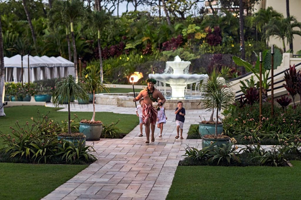 A man in traditional Hawaiin clothes holds a torch and leads some children through the grounds of Four Seasons Resort Maui at Wailea, one of the best hotels in Hawaii for a family vacation.
