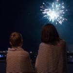 Two kids, wrapped in blankets, look up at Fourth of July fireworks.