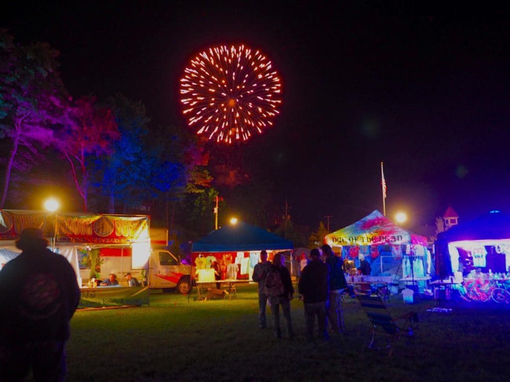 Several people mill about a neon-lit fair at night, while fireworks go off overhead in Lake George.