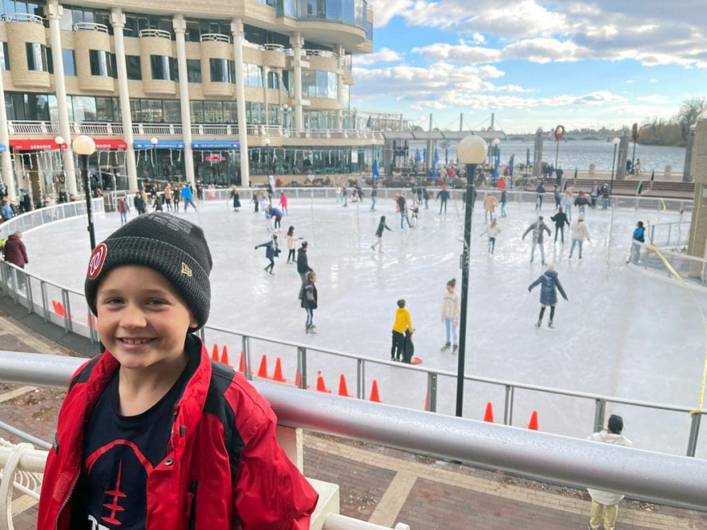 A young boy smiles, with a ice rink filled with skaters behind him, in the Georgetown neighborhood of Washington DC.