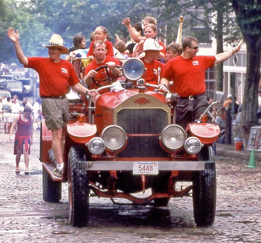 Several people dressed in red wave from a red, old-fashioned car during a Fourth of July parade in Nantucket.