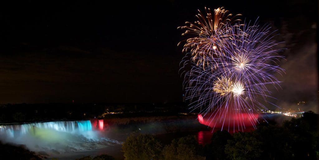 Niagara Falls lit up with special lights in hues of blue and green, while fireworks go off overhead.
