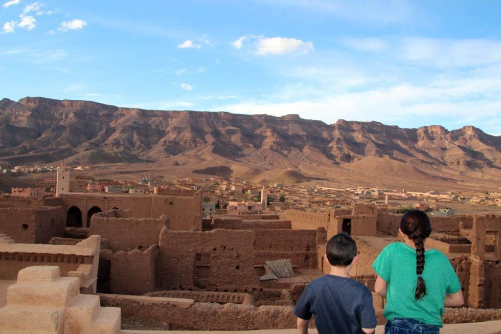 Two kids standing on the roof of a Moroccan riad look out onto the roofs of surrounding buildings and the desert in the distance.