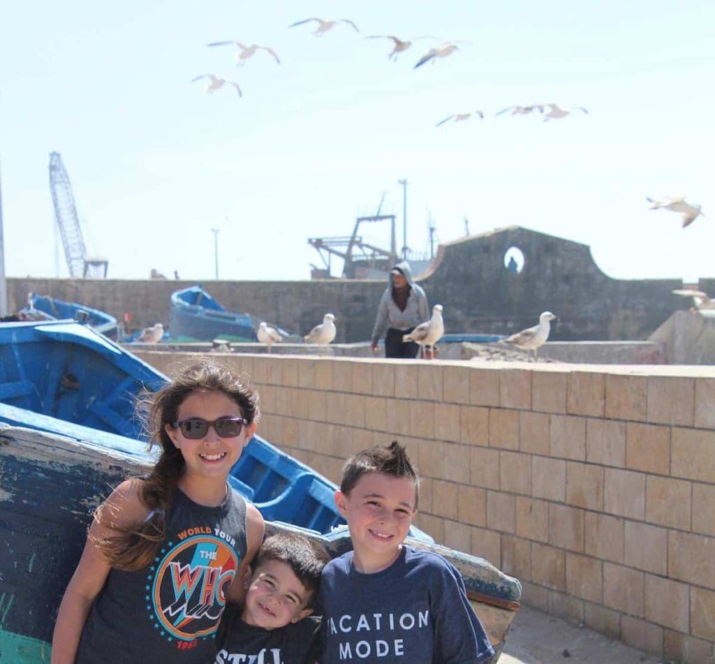 Three kids stand together with a shipping yard, seagulls, and crew members behind them.