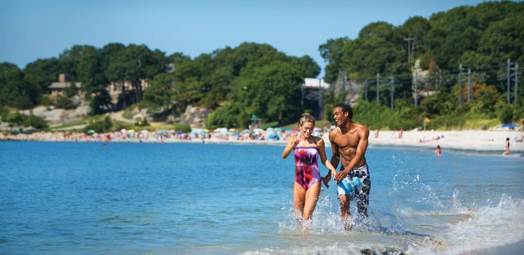 Two people splash through the shore of the lake within Rocky Neck State Park, with a crowded beach and line of trees in the distance.