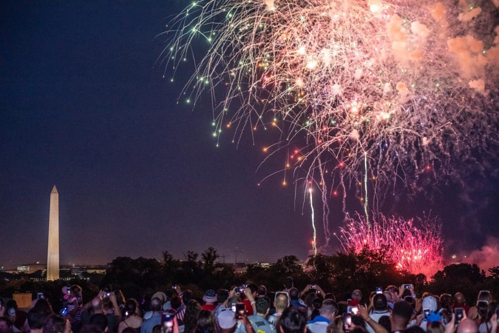 A huge crowd of people enjoy fire works, while the Washington Monument can be seen in the distance.