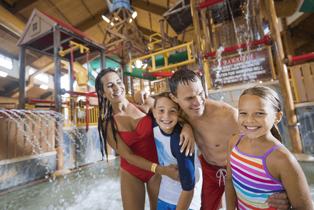 A family of four, all wearing bathing suits, stand together, smiling, while enjoying the indoor water park area at Wilderness Resort.