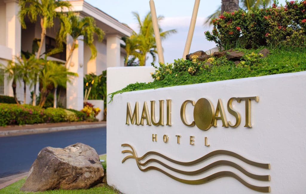 The chic entrance sign for Maui Coast Hotel, with gold lettering agains a white background, and the entrance to the resort in the background.