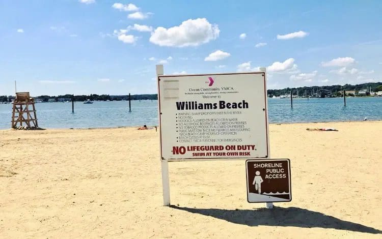 A close up of the safety sign at Williams Beach, near Mystic, with a sandy shore and water in the distance on a sunny day.