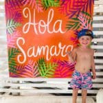 A young boy wearing a swim suit and hat stands next to a vibrant sign reading "Hola Samara".