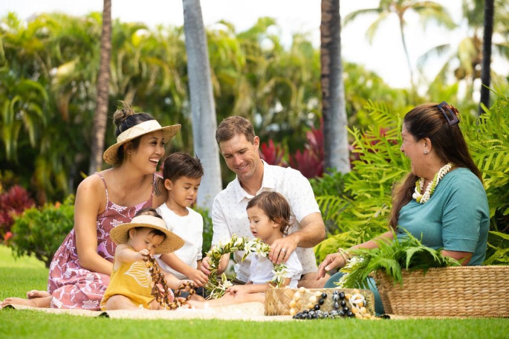 A family of four sits with a hotel staff member at Fairmont Orchid, Hawaii, while enjoying a picnic amongst lush greenery.