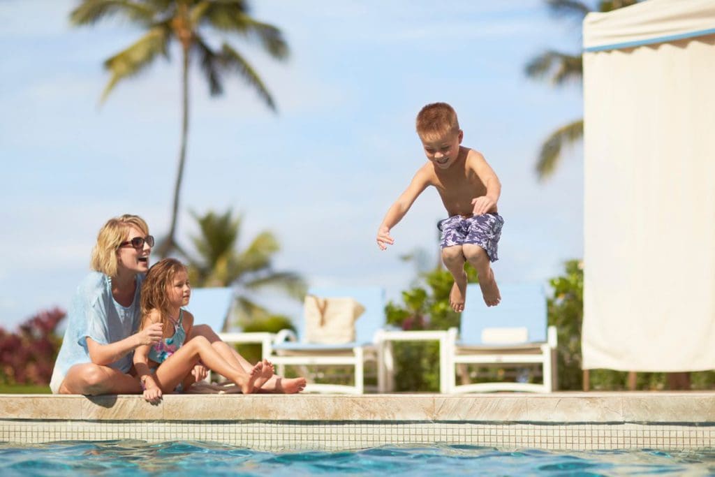 A mom and her young child sit at the edge of the pool, while an older child jumps into the pool at Four Seasons Resort Maui at Wailea.
