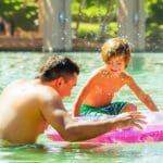 A father and son play together in a pool, using a large pink flotation device.