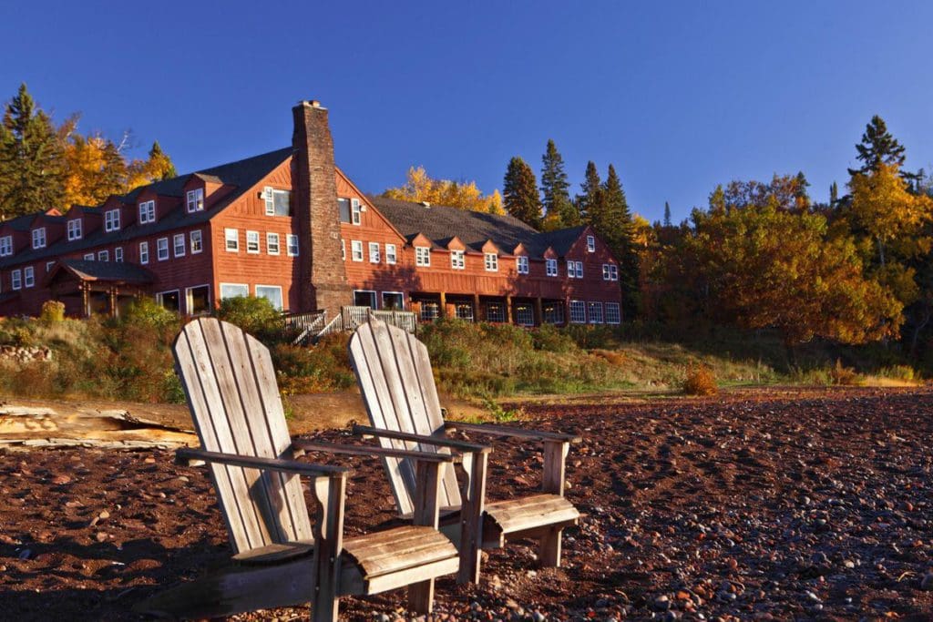 Two Adirondack chairs sit on the pebbly shore of Lake Superior with Lutsen Resort in the background.