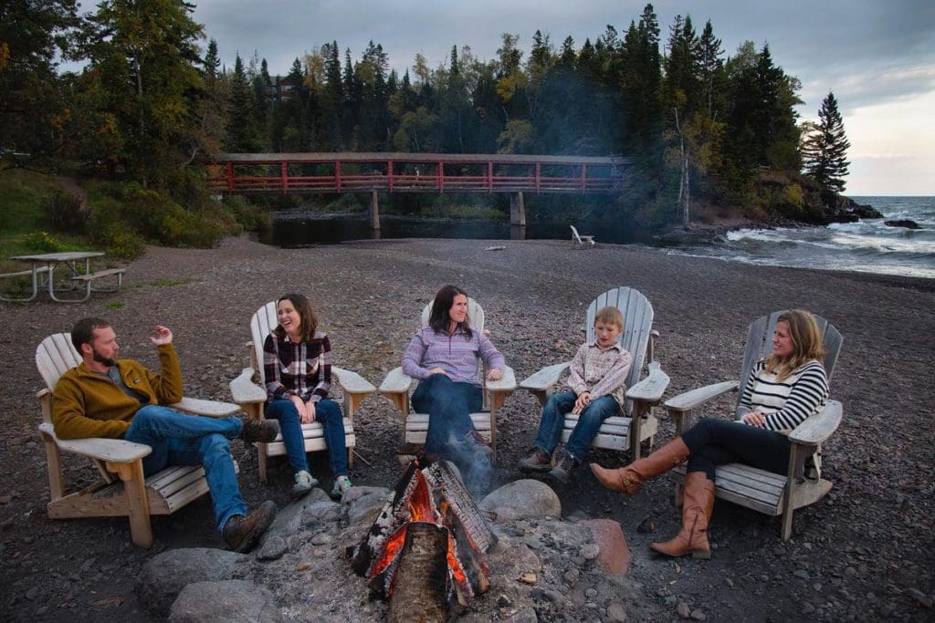 A family of five sits within Adirondack chairs around a beach fire on the shore of Lake Superior, while staying at Lutsen Resort, one of the best summer resorts in Minnesota for families.