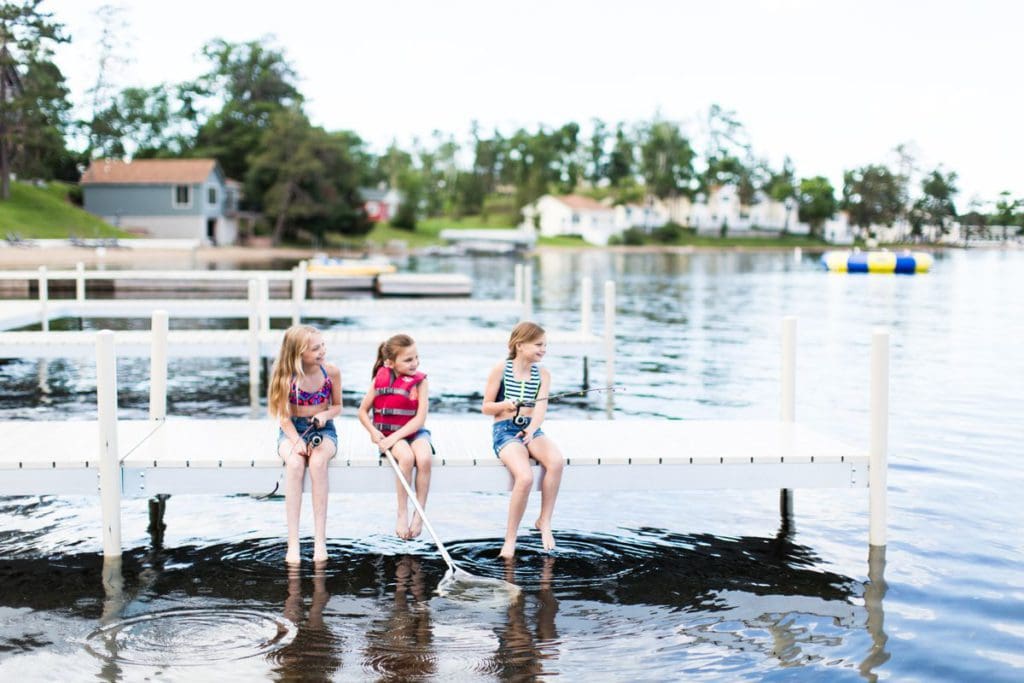 Three young girls, wearing life jackets, sit on the edge of a dock, dangling their feet into the water on a summer day at Maddens on Gull Lake.