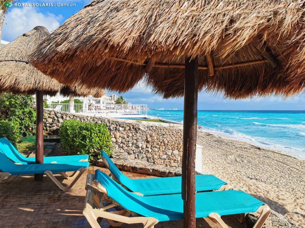Two blue beach loungers under a grass cabana sit on the beach looking at the ocean at Royal Solaris Cancun.