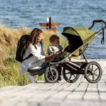 A mom kneels by her young son next to a Thule Urban Glide 2 stroller, with an ocean and tall beach grass in the distance.
