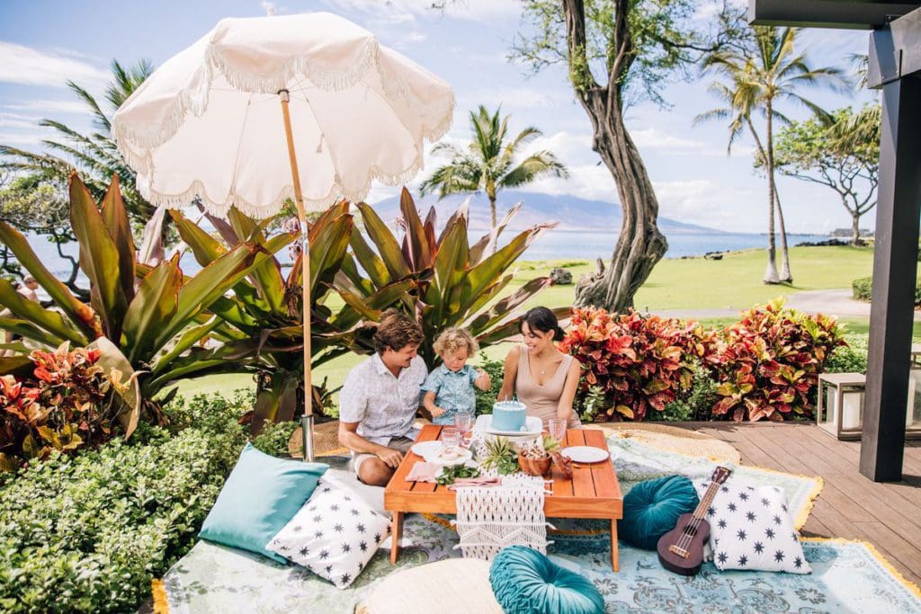 A family of three sits together enjoying a picnic at the luxurious Wailea Beach Resort in Maui, one of the best hotels in Hawaii for a family vacation.