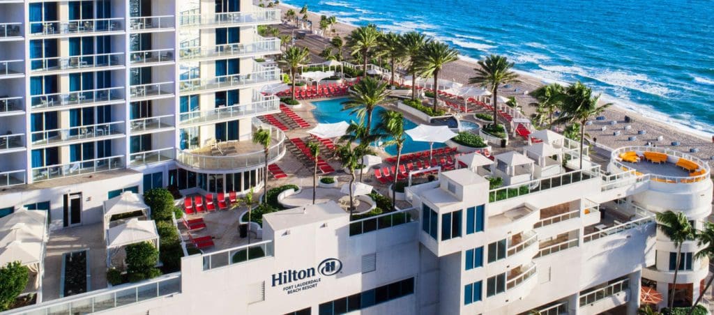 An aerial view of the roof top pool and terrace at Hilton Fort Lauderdale Beach Resort, with large resort buildings nearby, and the ocean in the distance.