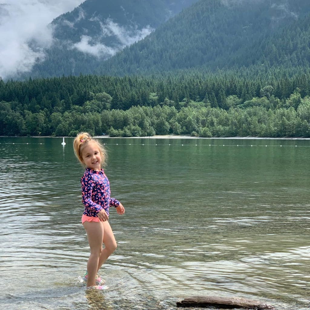 A young girl wearing a swimming suit stands on the shores of the lake within Golden Ears Provincial Park in British Colombia, one of the best vacation destinations for families In Canada.