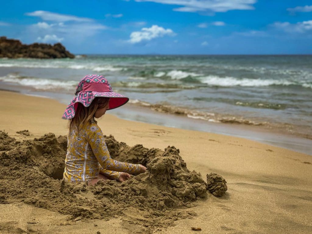 A young girl sits in a hole she dug in the stand, while enjoying a sunny day on Colón Park Beach near the water.