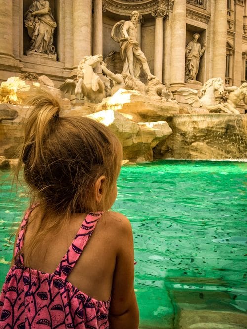 A young girl leans over the side of the Trevi Fountain to peer into the water, a must stop on our Rome itinerary with kids.