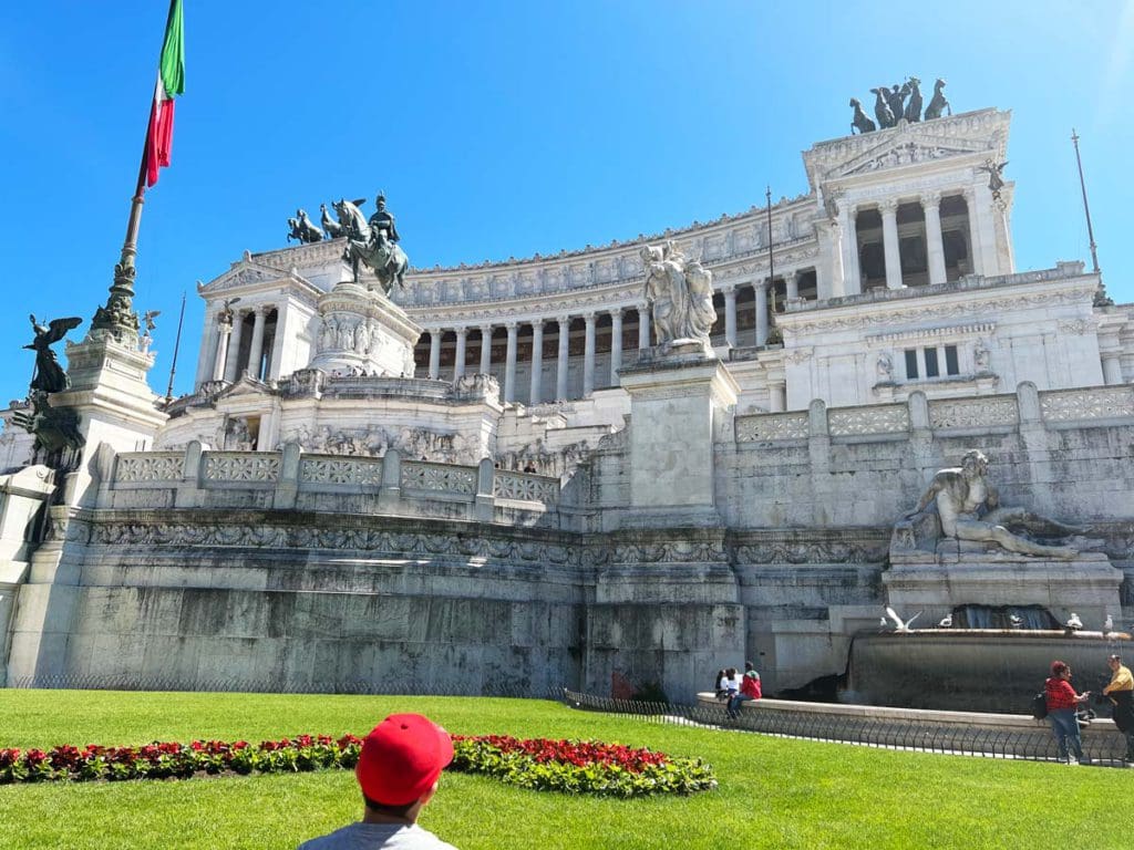 A young boy, wearing a red baseball hat, looks up at the historic Capitoline Hill on a sunny day, an optional stop on our Rome itinerary with kids.