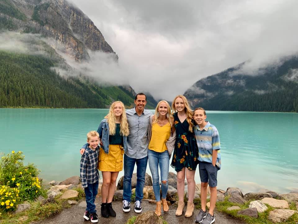 A large family stands together, smiling, in front of the turquoise waters of Moraine Lake in Canada.