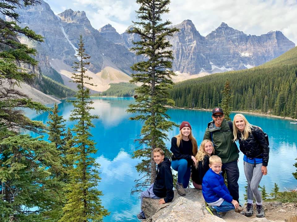 A large family stands together on a cliffside overlooking Moraine Lake in Alberta, with large pine trees near them.