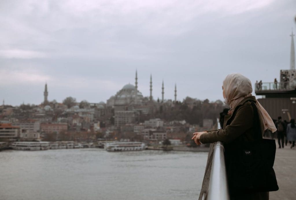 A woman wearing a hijab leans over a railing along the water while looking at the Istanbul skyline.