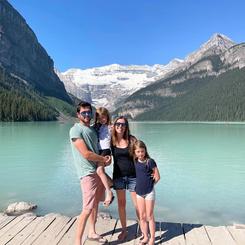 A family of four stands together, smiling, on a lakeside dock at Lake Louise in Alberta.